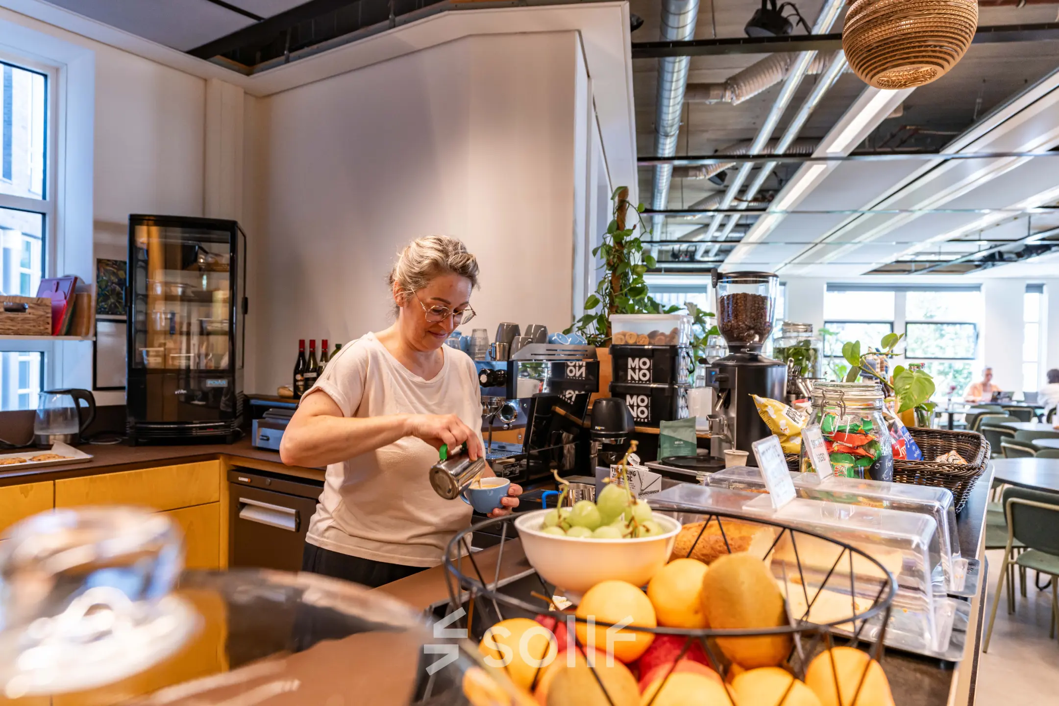 Binnenzicht van kantoorruimte op Fluwelen Burgwal 58, Den Haag Centrum, met een persoon die koffie zet bij een pantry, omringd door fruit en planten.
