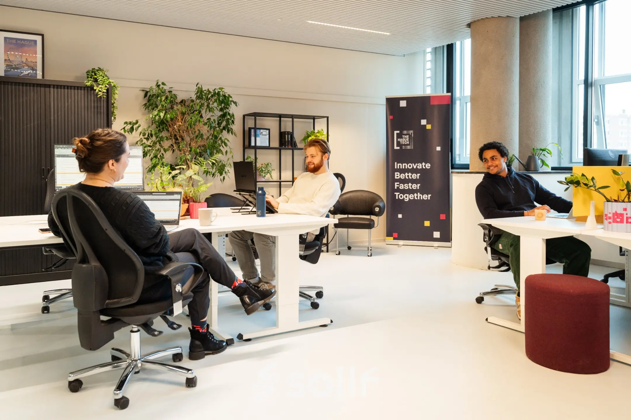 Zakelijke omgeving op Waldopstraat 5, Den Haag Centrum, met mensen in gesprek in een moderne kantoorruimte. De ruimte is voorzien van bureaus en enkele planten.