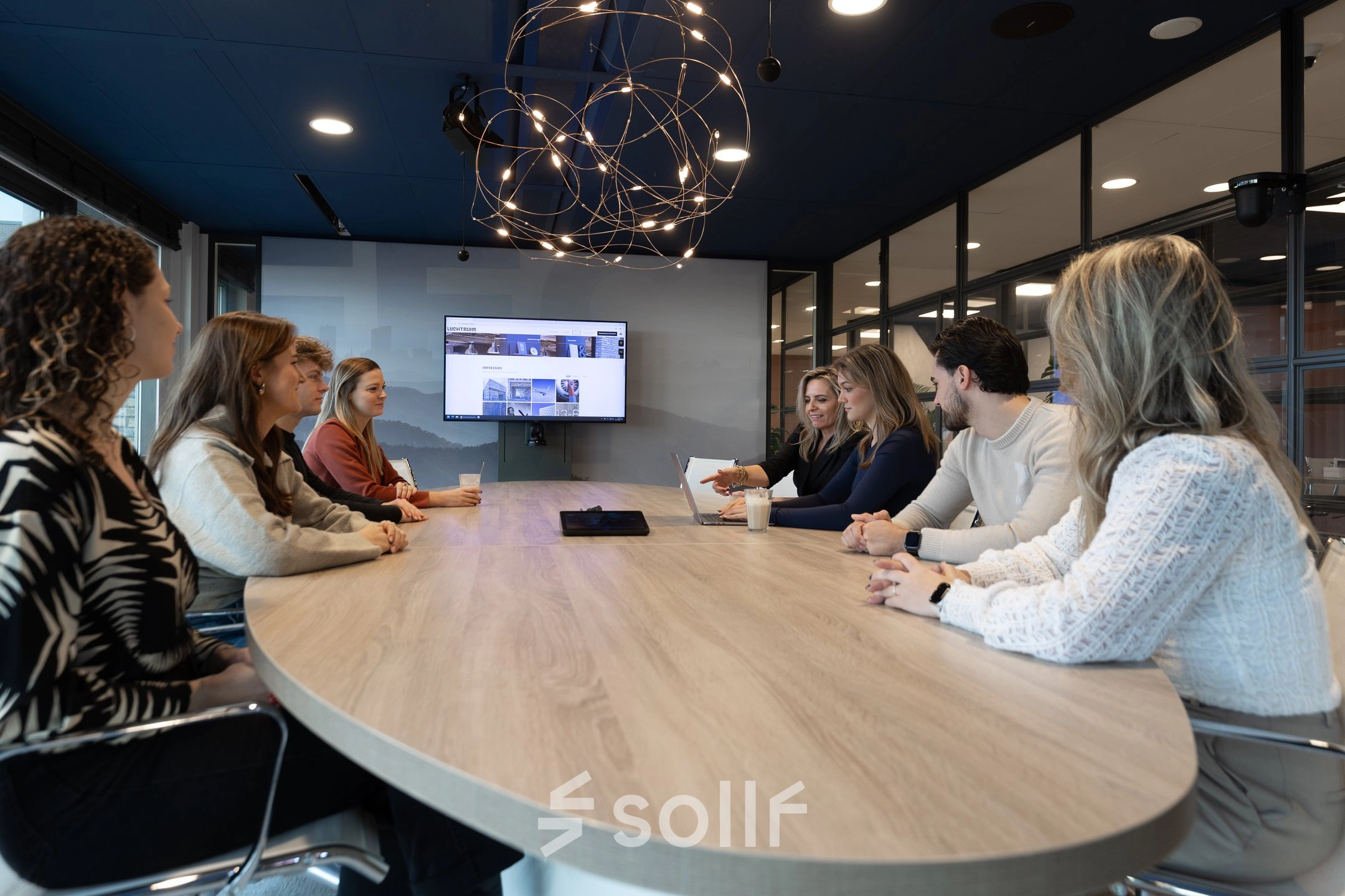 Zakelijk overleg in een modern kantoorpand op Flight Forum 120-159, Eindhoven Airport. Zes personen zijn in gesprek rond een houten tafel, met een groot scherm op de achtergrond. Ideaal voor kantoorruimte huren.