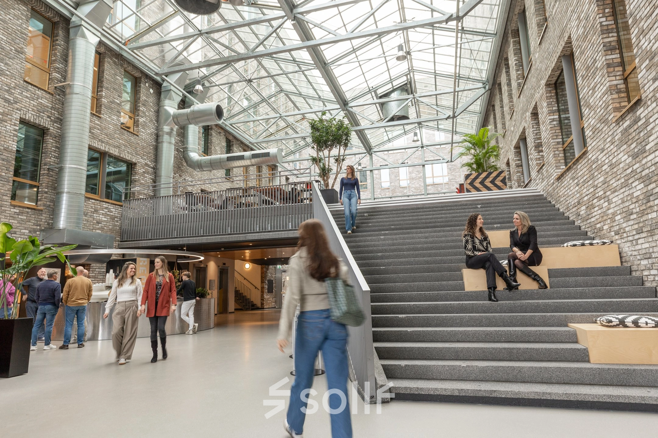 Moderne kantoorruimte met glazen dak op Flight Forum 120-159 in Eindhoven Airport, waar mensen werken en socializen op verschillende niveaus. Ideaal om kantoorruimte te huren in een inspirerende omgeving.