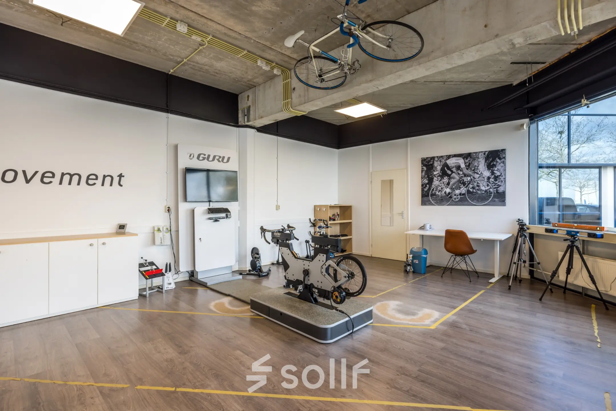 Kantoorruimte te huur op Luchthavenweg 81, Eindhoven Airport. De ruimte heeft een modern interieur met houten vloeren en is deels gemeubileerd. Inclusief werktafel, fietsensimulator en een fiets aan het plafond.