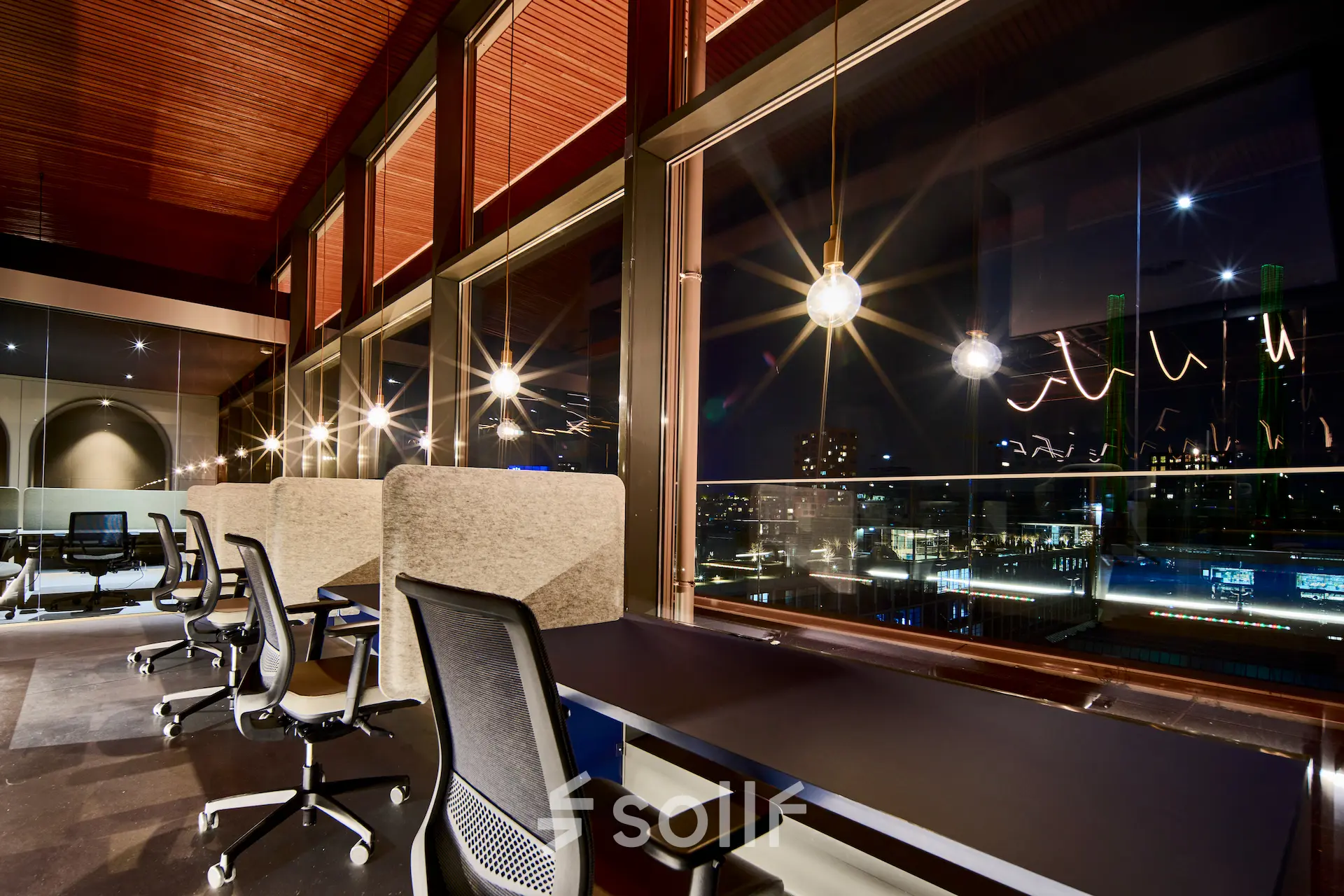 Moderne kantoorruimte met uitzicht op de nachtelijke skyline van Eindhoven, gedeeltelijk gemeubileerd met bureaus en stoelen. Perfect voor wie kantoorruimte wil huren. Gelegen op Achtseweg Zuid 159R, in Strijp-S.