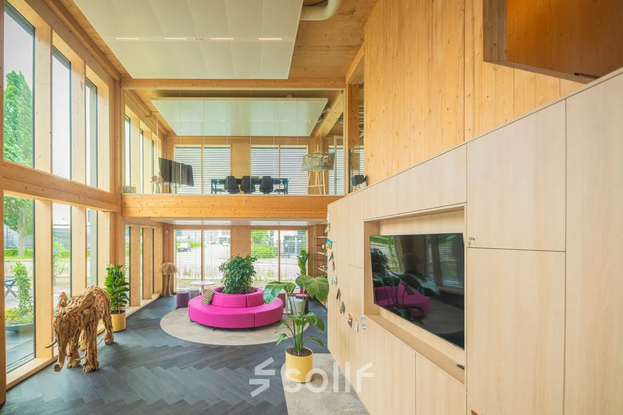 Lichte en moderne kantoorruimte met houten accenten en grote ramen op Moutlaan 1-23, Enschede. Roze bank en planten verlevendigen het interieur. Ideaal voor werk en vergaderingen.