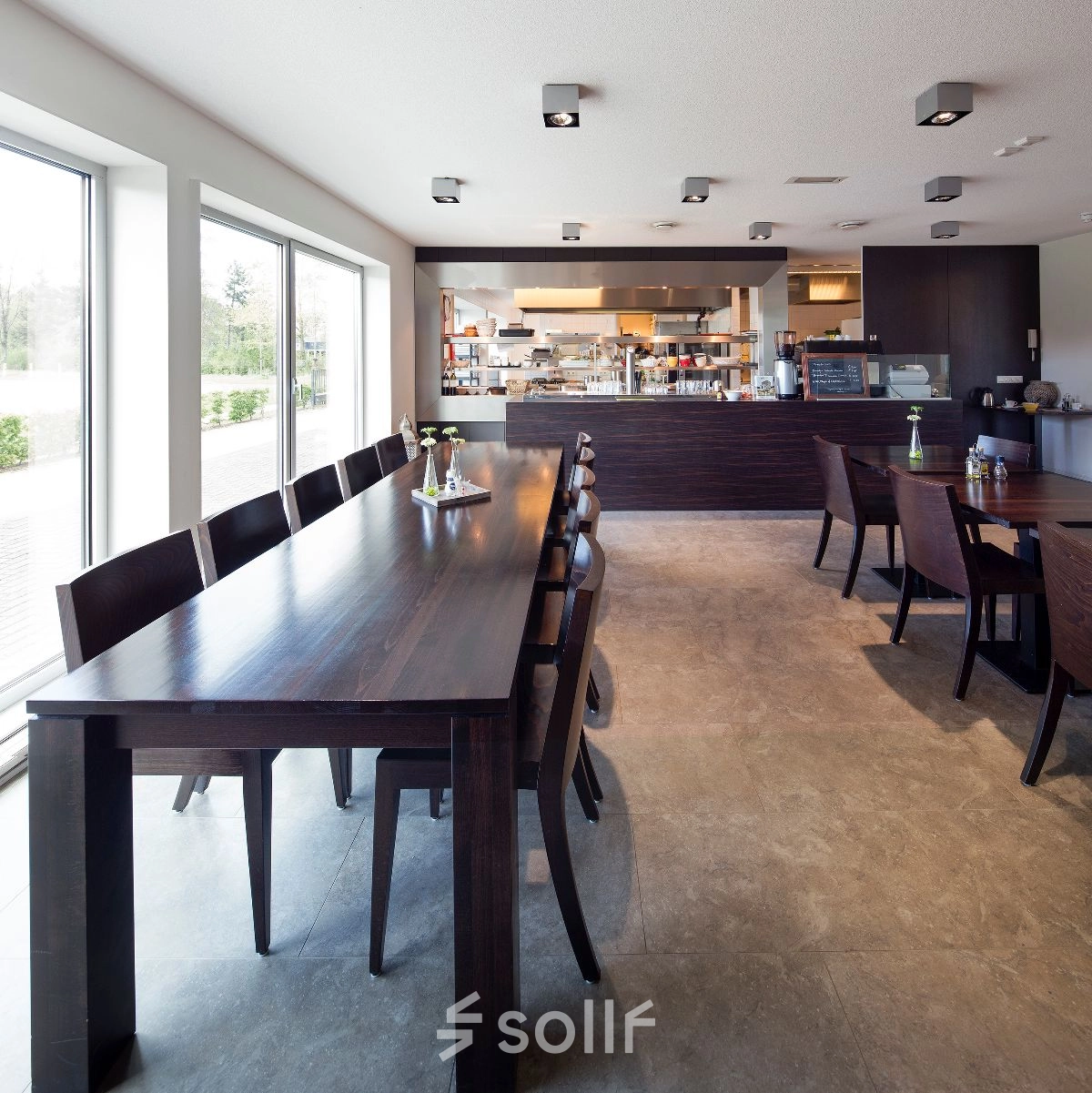 Moderne kantoorruimte te huur in Geleen met ruime tafels en een open keuken. Het interieur is gemeubileerd met hoogwaardige, donkere houten meubels. Groot raam biedt veel natuurlijk licht.