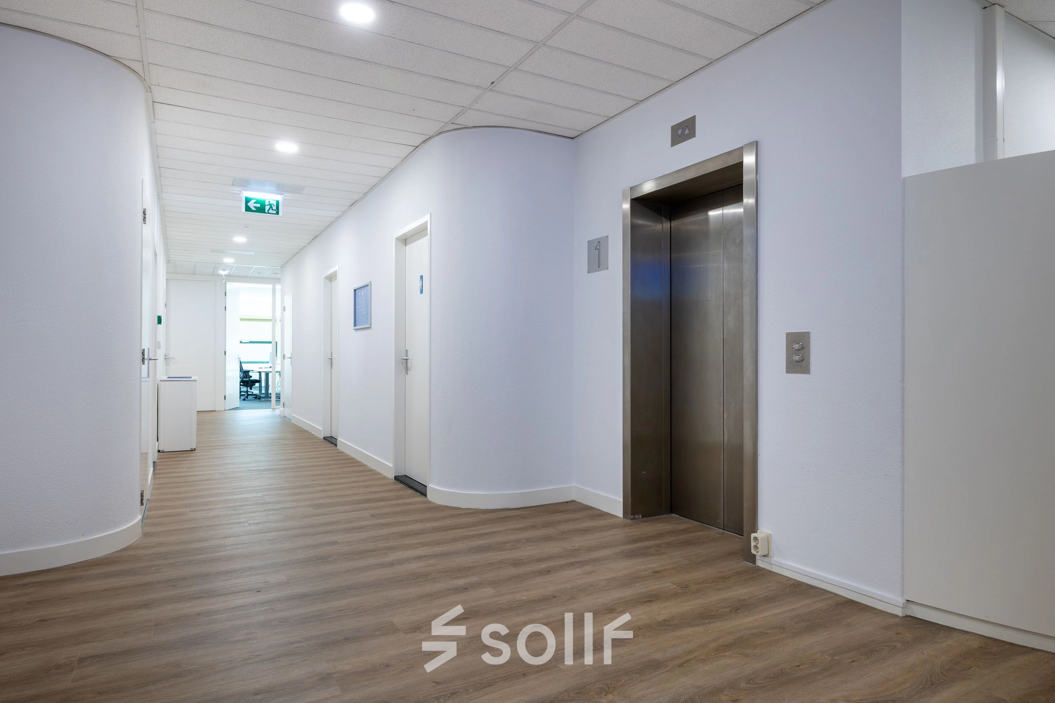 Een lichte gang in een kantoorpand met een houten vloer, witte muren en een moderne lift. De ruimte is goed verlicht en leidt naar verschillende kantoorruimtes.