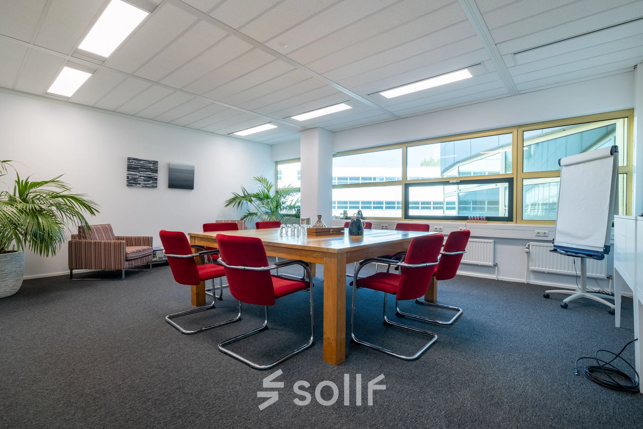 Ruime vergaderkamer in kantoorpand aan Groningenweg 8 8, Gouda, met rode stoelen rondom een houten tafel, grote ramen en kamersplanten. Ideaal voor kantoorruimte huren.