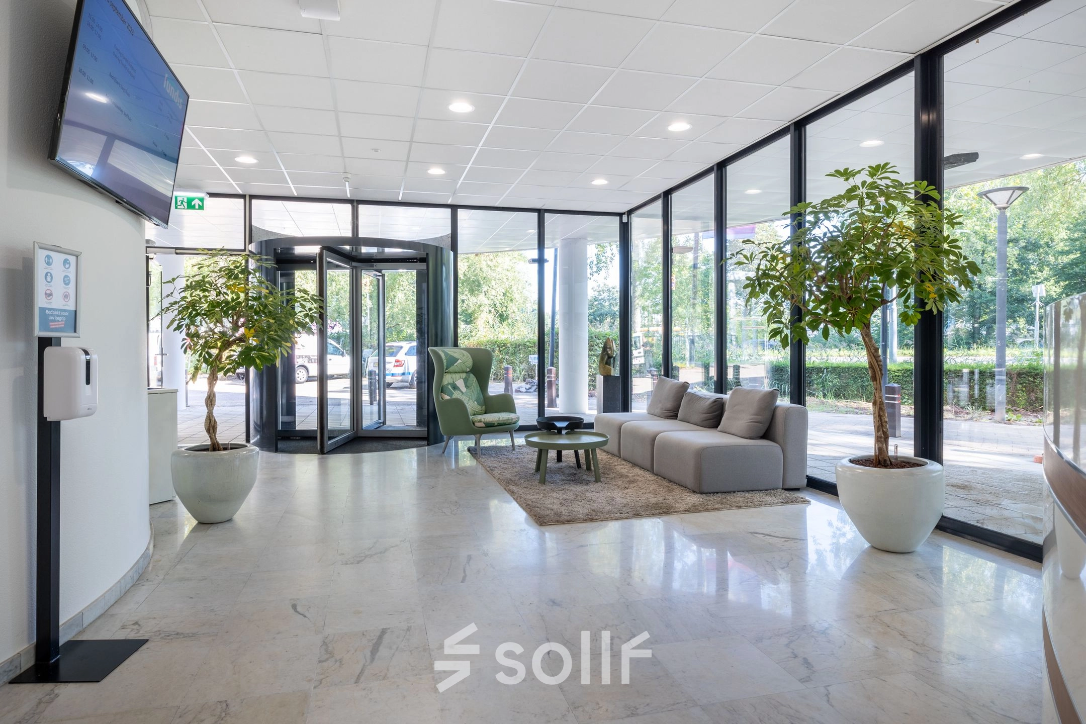 Lichte entree van een kantoorpand in Gouda met glazen deuren, een comfortabele bank, en planten. Ideaal voor kantoorruimte huren.