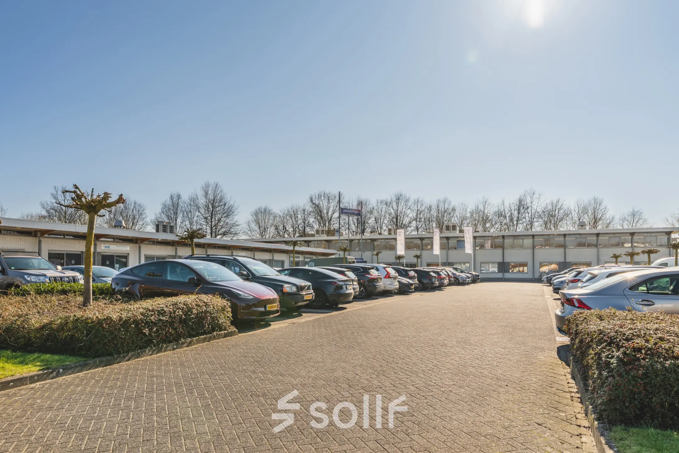 Parkeerplaats voor een kantoorpand aan de Steenovenweg 5 in Helmond, omringd door geparkeerde auto's en groene heggen onder een heldere blauwe lucht.