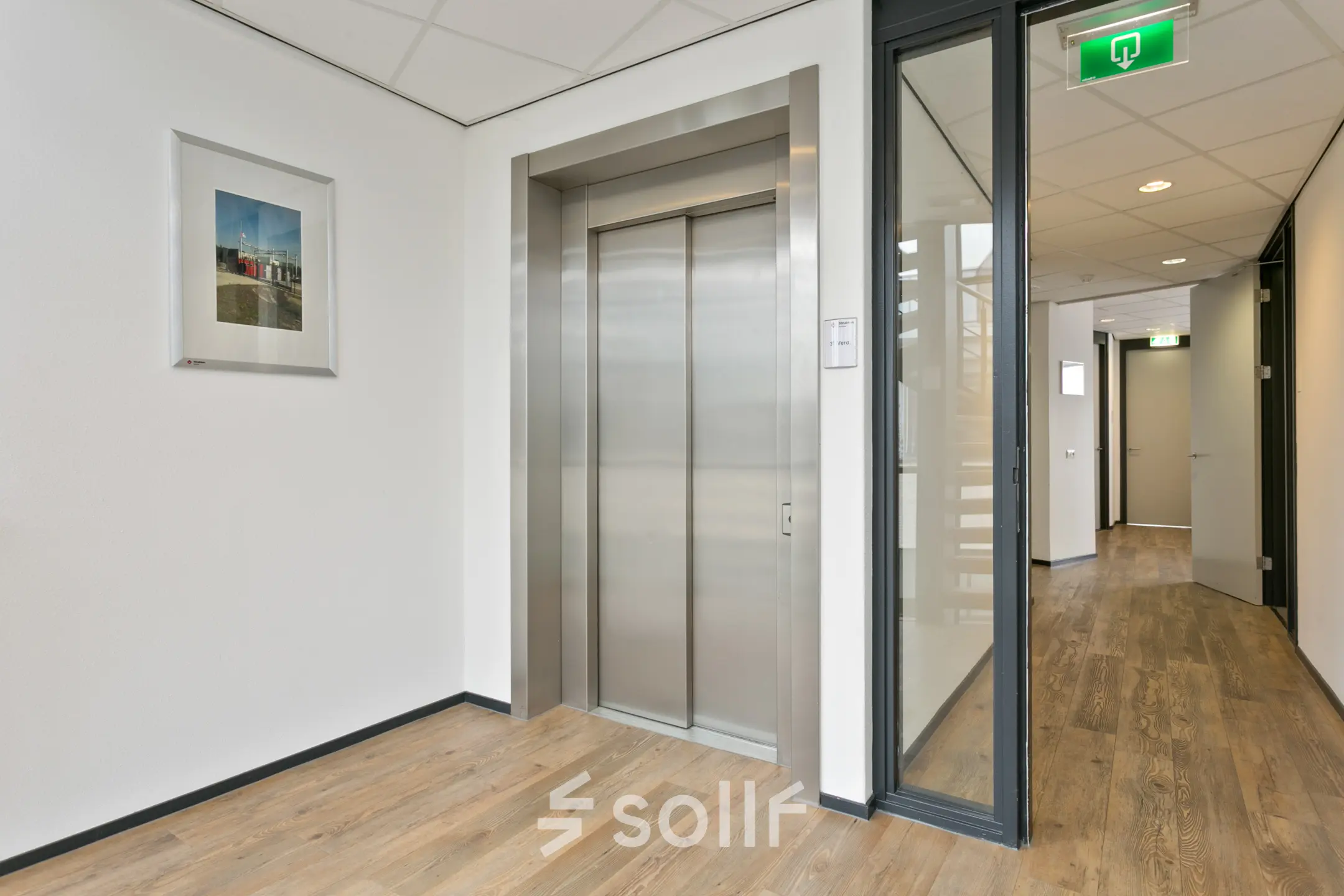Kantoorruimte huren in Hengelo aan de Welbergweg 60 met nette entree met lift en houten vloer. Het licht binnendringt door een glazen wand, terwijl kunst aan de muur zorgt voor een stijlvolle touch.