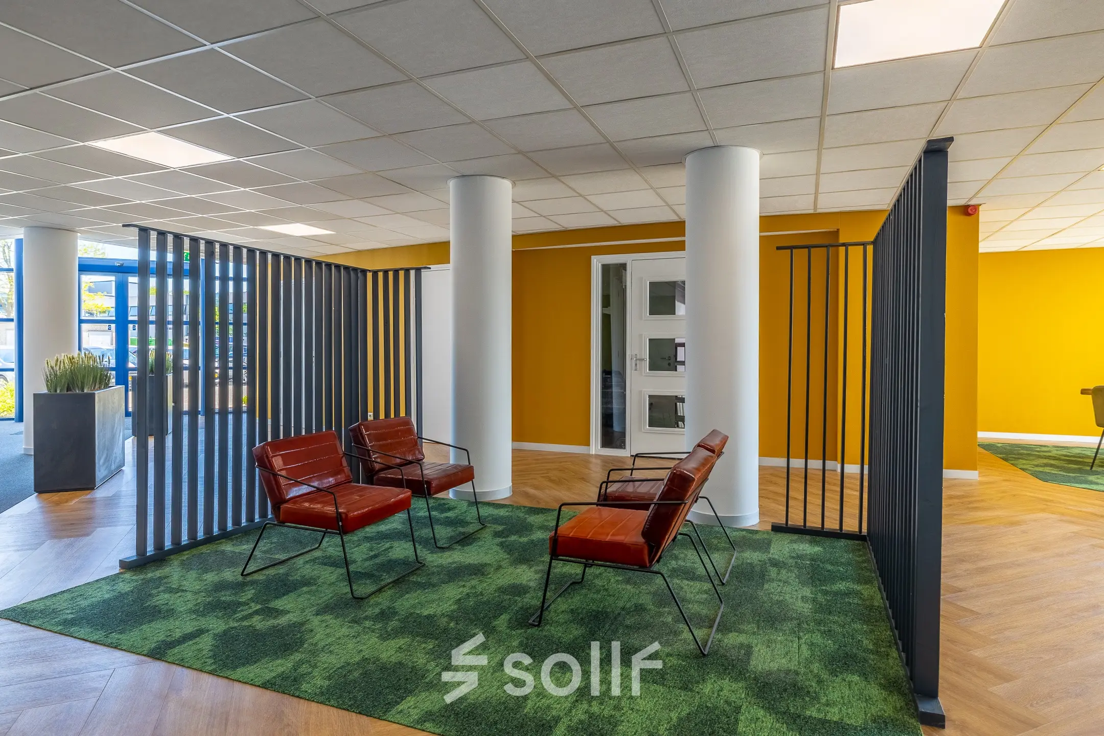 Moderne kantoorruimte met kleurrijke muren en comfortabele zithoek, ideaal voor informele vergaderingen of pauzes. Glaswand zorgt voor veel licht en een uitnodigende sfeer om te werken.