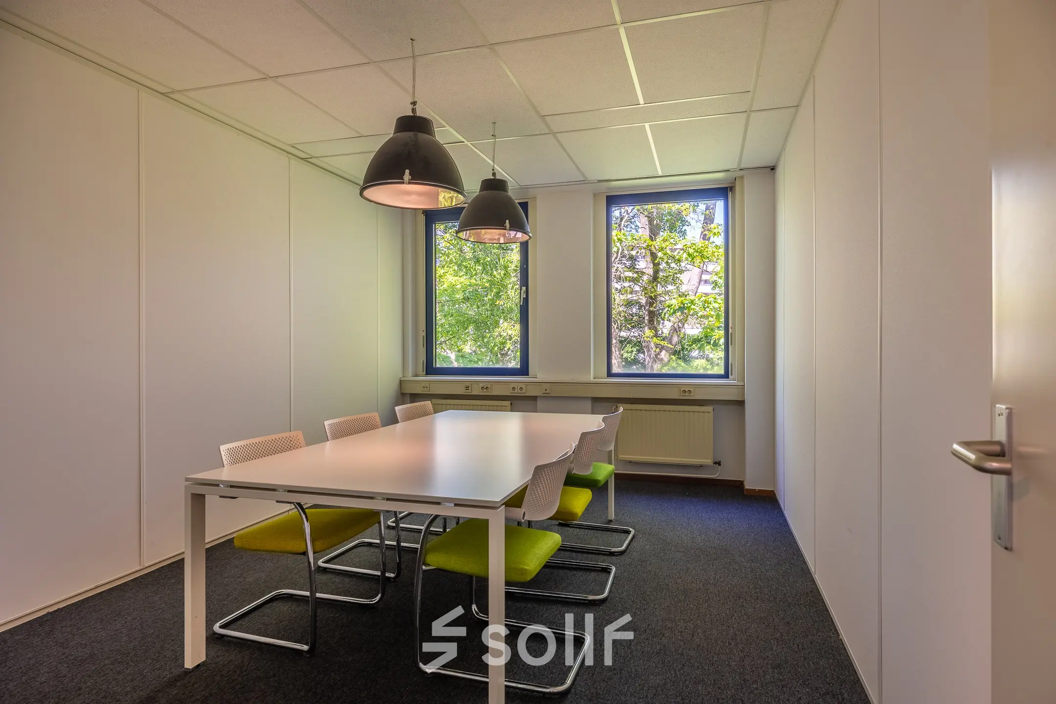 Kantoorruimte te huur aan de Franciscusweg 219 in Hilversum. Het interieur heeft een vergadertafel met stoelen, grote ramen geven zicht op groen buiten.