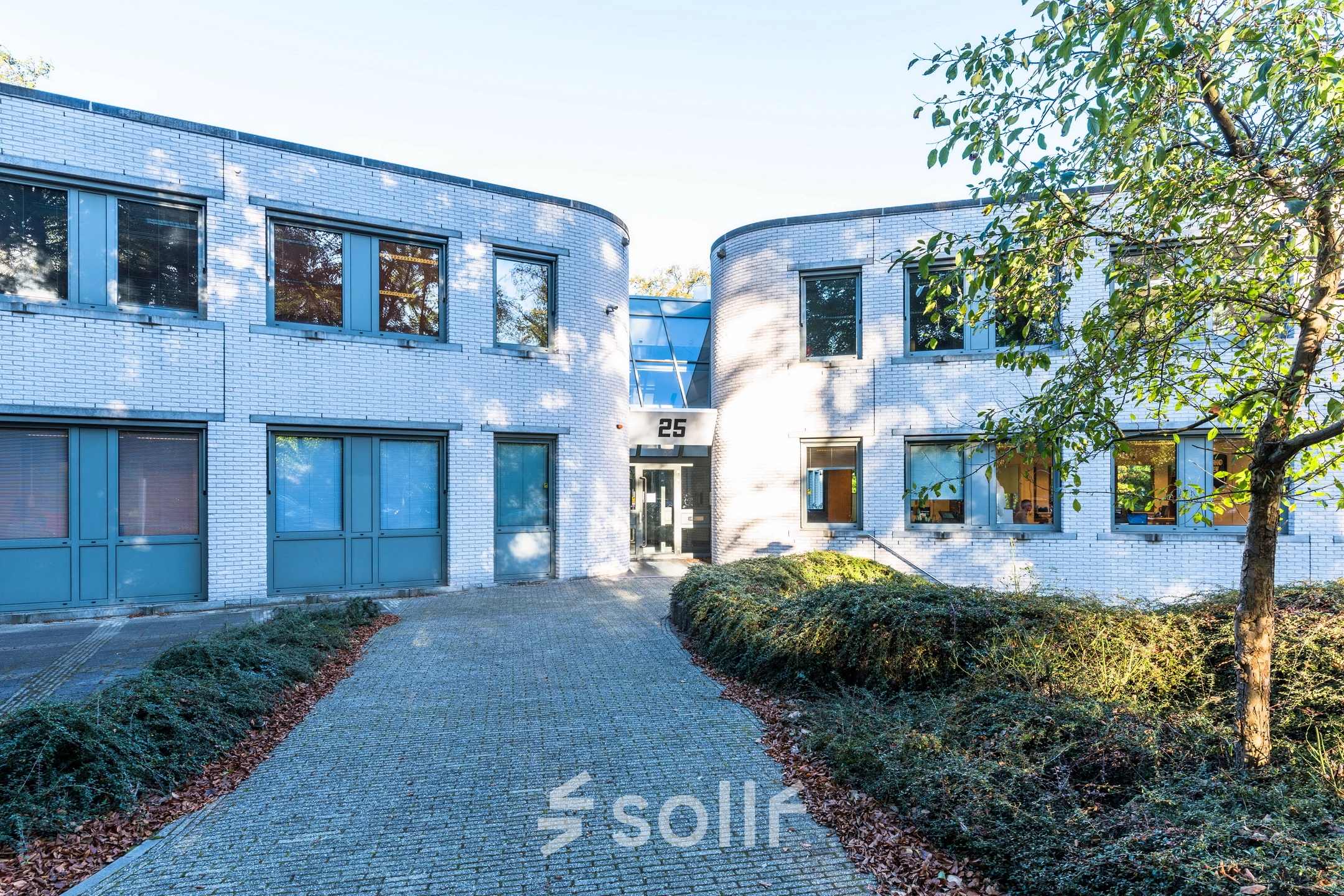Uitzicht op de buitenkant van het kantoorpand op Mozartlaan 25 in Hilversum, geschikt voor kantoorruimte huren. Het entreepad leidt naar een moderne glazen ingang die omringd is door witte bakstenen muren.
