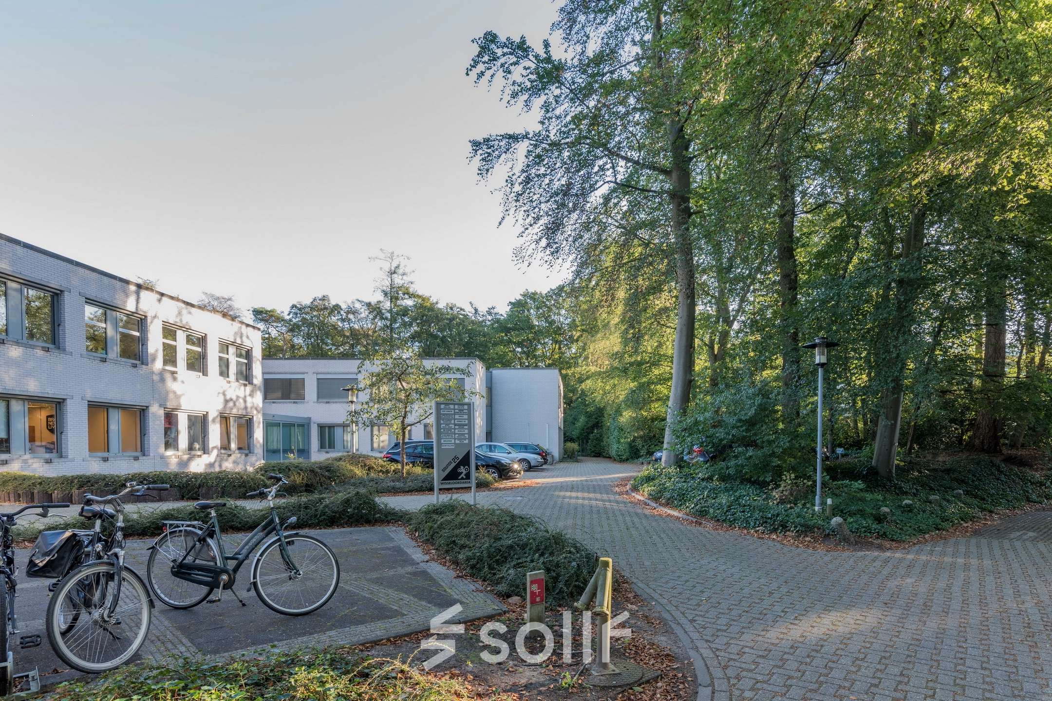 Kantoorpand aan de Mozartlaan 25 in Hilversum, omgeven door bosrijke omgeving. Parkeerplaats met fietsen en auto's zichtbaar.