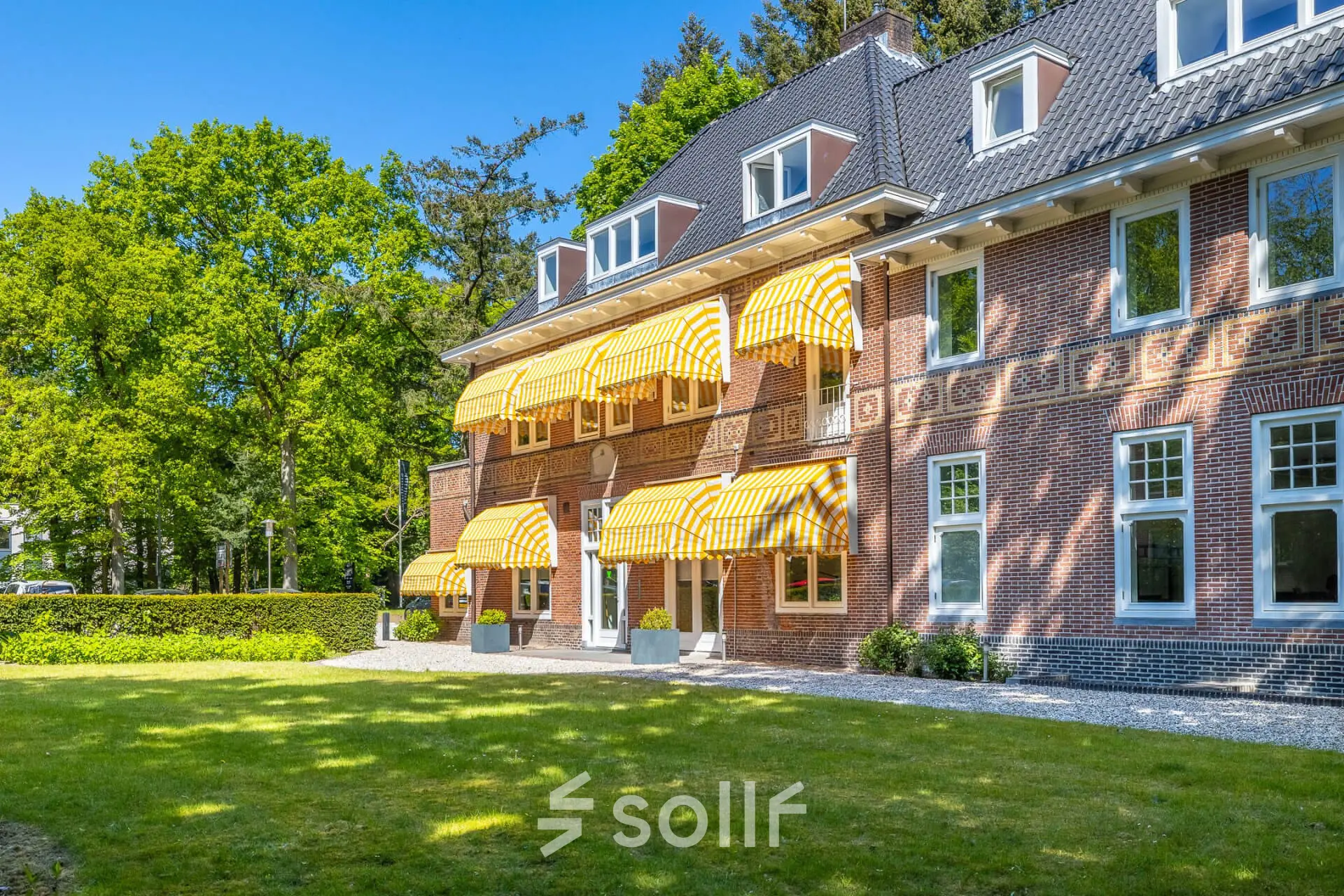 Klassiek kantoorpand aan de Utrechtseweg 47 in Hilversum, omgeven door groene beplanting en uitgerust met gele zonwering. Ideaal voor wie kantoorruimte huren wil in een inspirerende omgeving.