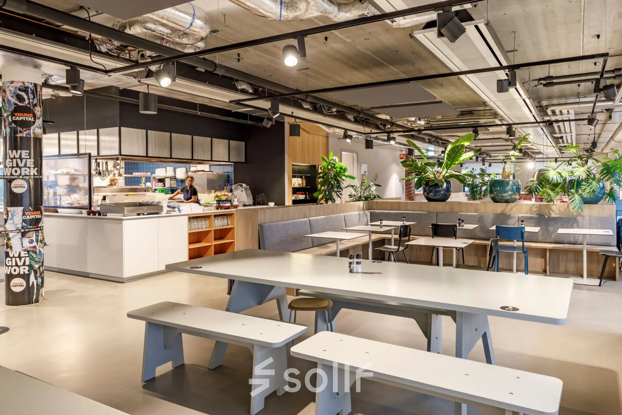 Moderne kantoorruimte in Hoofddorp met gemeubileerde werkplekken, een open keuken en groene accenten. Mensen zien bezig met het bereiden van eten. Ideaal om een kantoorruimte te huren.