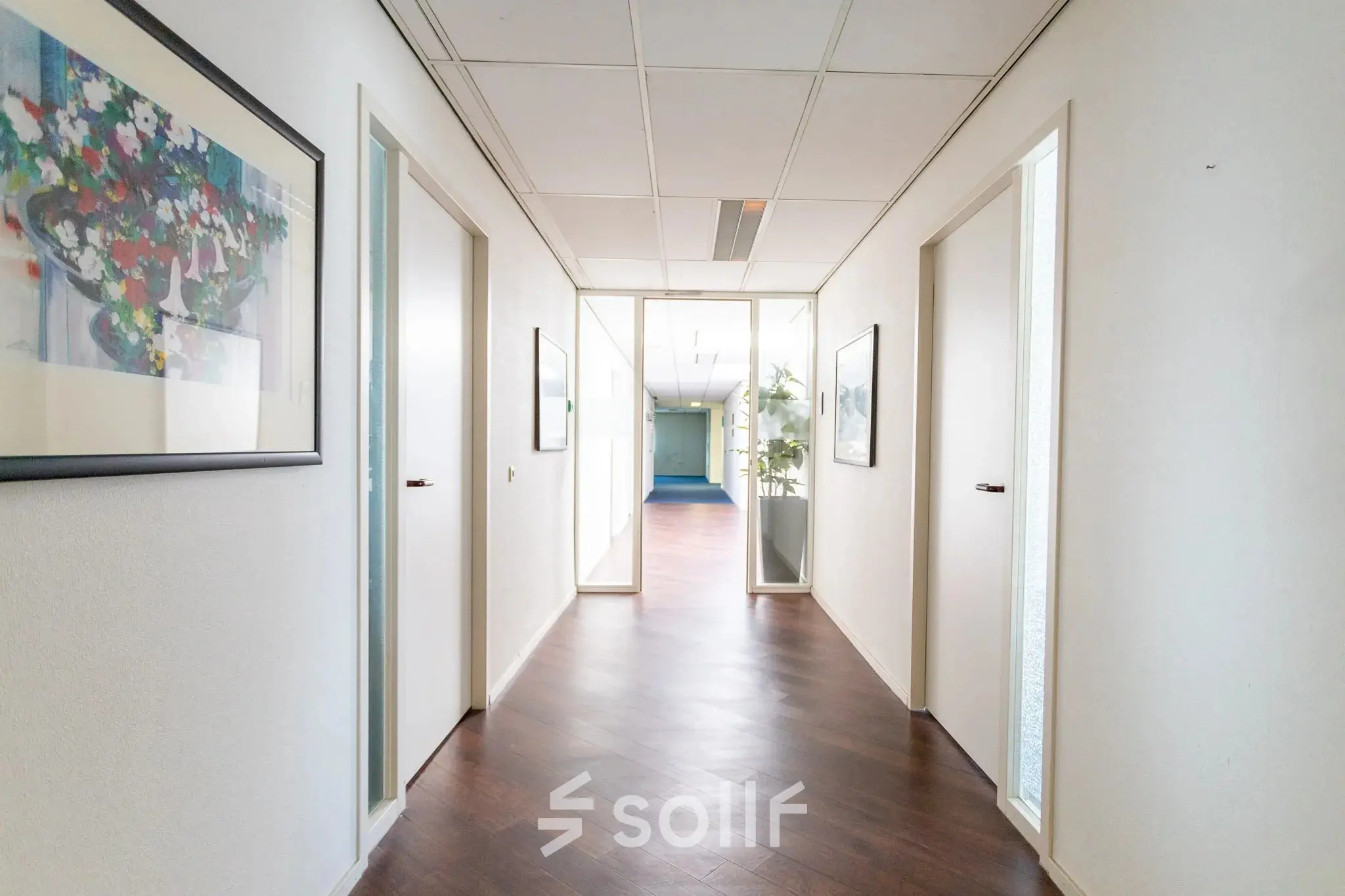 Heldere gang in een kantoorpand op Sandtlaan 36, Katwijk, met houten vloer en kunst aan de muur. Ideaal voor bedrijven die kantoorruimte willen huren met ruimte voor creativiteit en productiviteit.