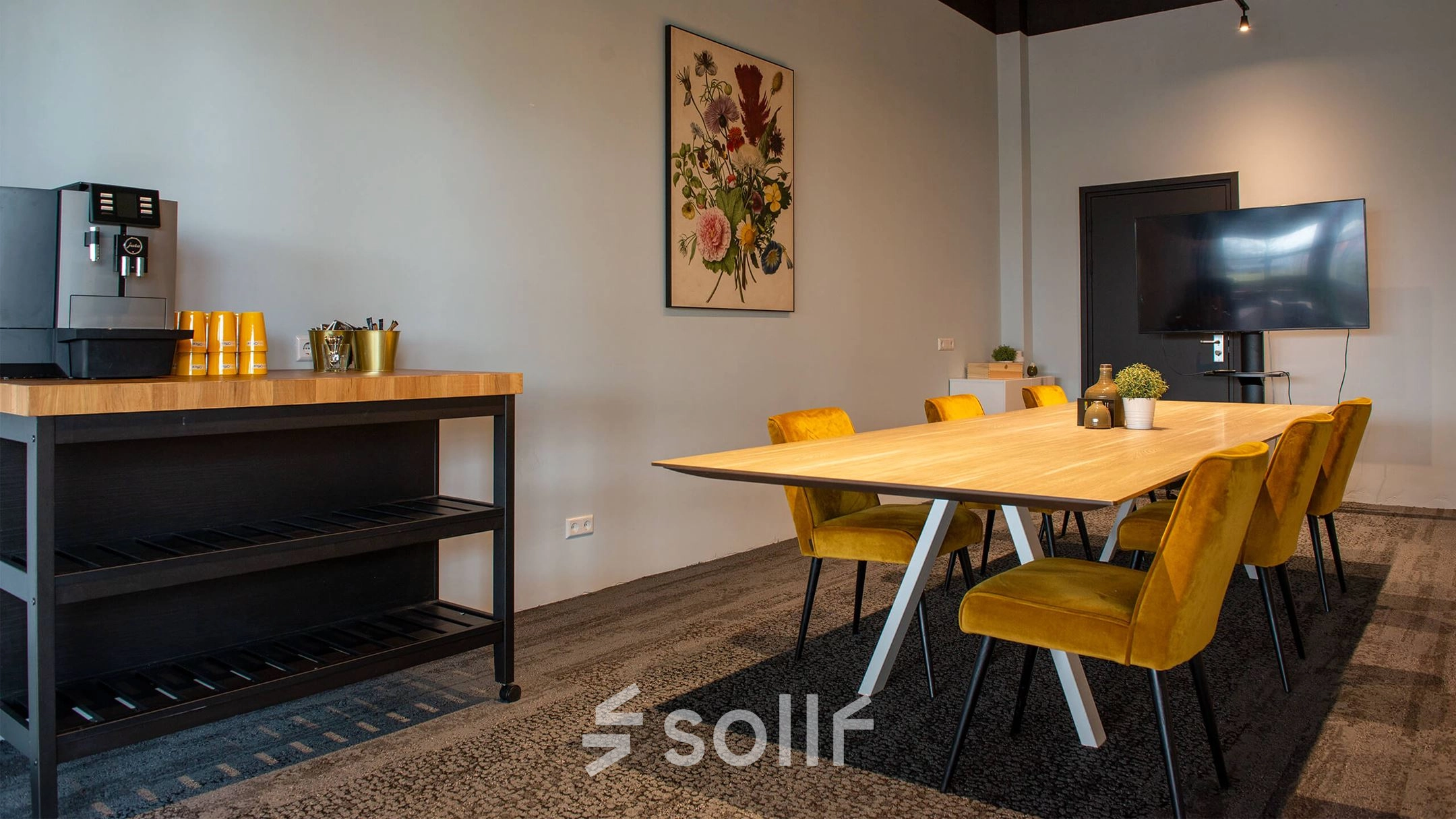 Moderne kantoorruimte in Meppel met vergadertafel en felgekleurde stoelen, ingericht om professioneel te huren. Er is een koffiezetapparaat en een beeldscherm aan de muur.