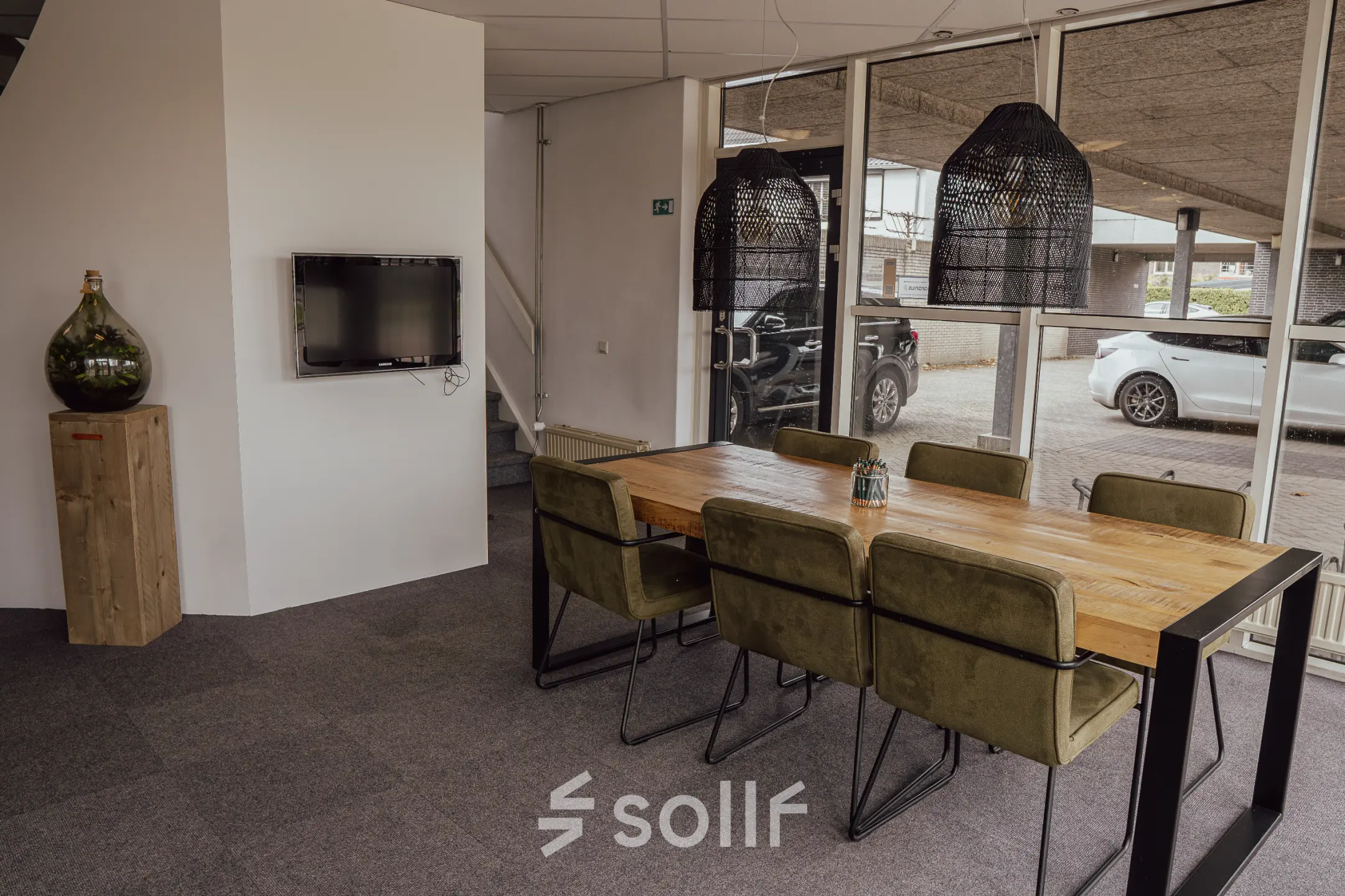 Kantoorruimte huren in Oosterhout Nb, Abdis van Thornstraat 74. Modern kantoorpandinterieur met houten tafel en groene stoelen, tegen een glazen wand met uitzicht op buiten. Neutrale tinten en stijlvolle verlichting.