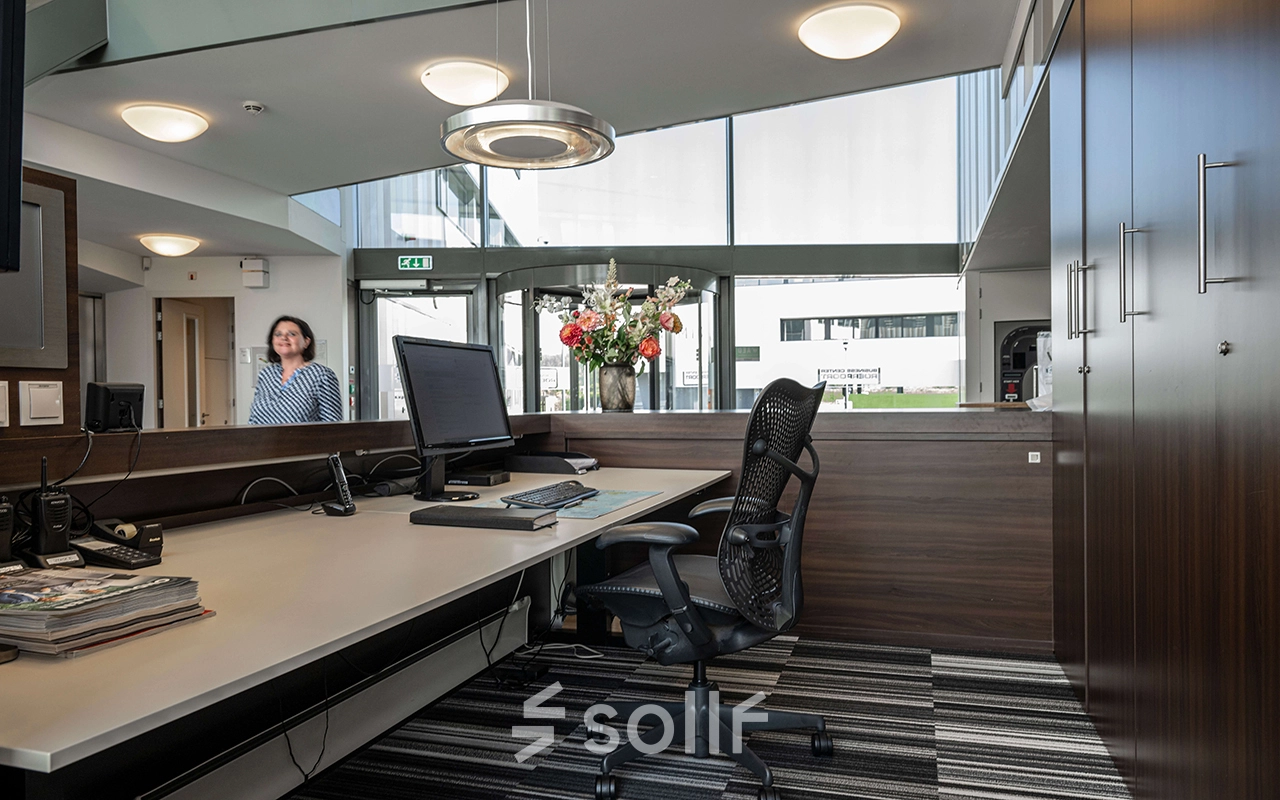 Receptieruimte in een kantoorpand aan Boven de Wolfskuil 3, Roermond, met balie, computerscherm en stoelen. Een persoon staat binnen te kijken vanuit de ingang.