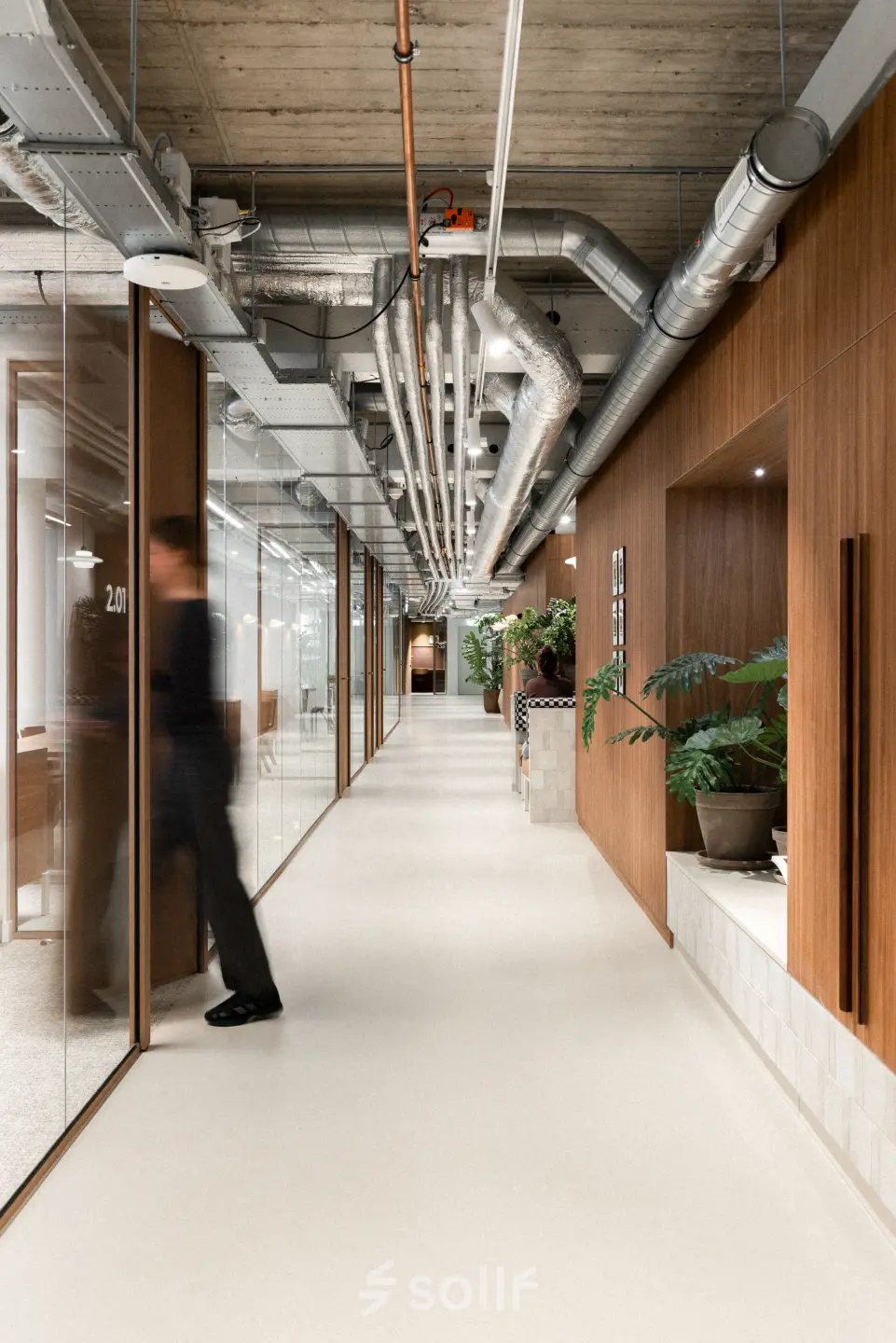 Moderne kantoorruimte te huur op Stadhuisplein 9-23 in Rotterdam Centrum, met glazen wanden, leidingen aan het plafond en groene planten. Een persoon loopt door de gang richting een kantoor.