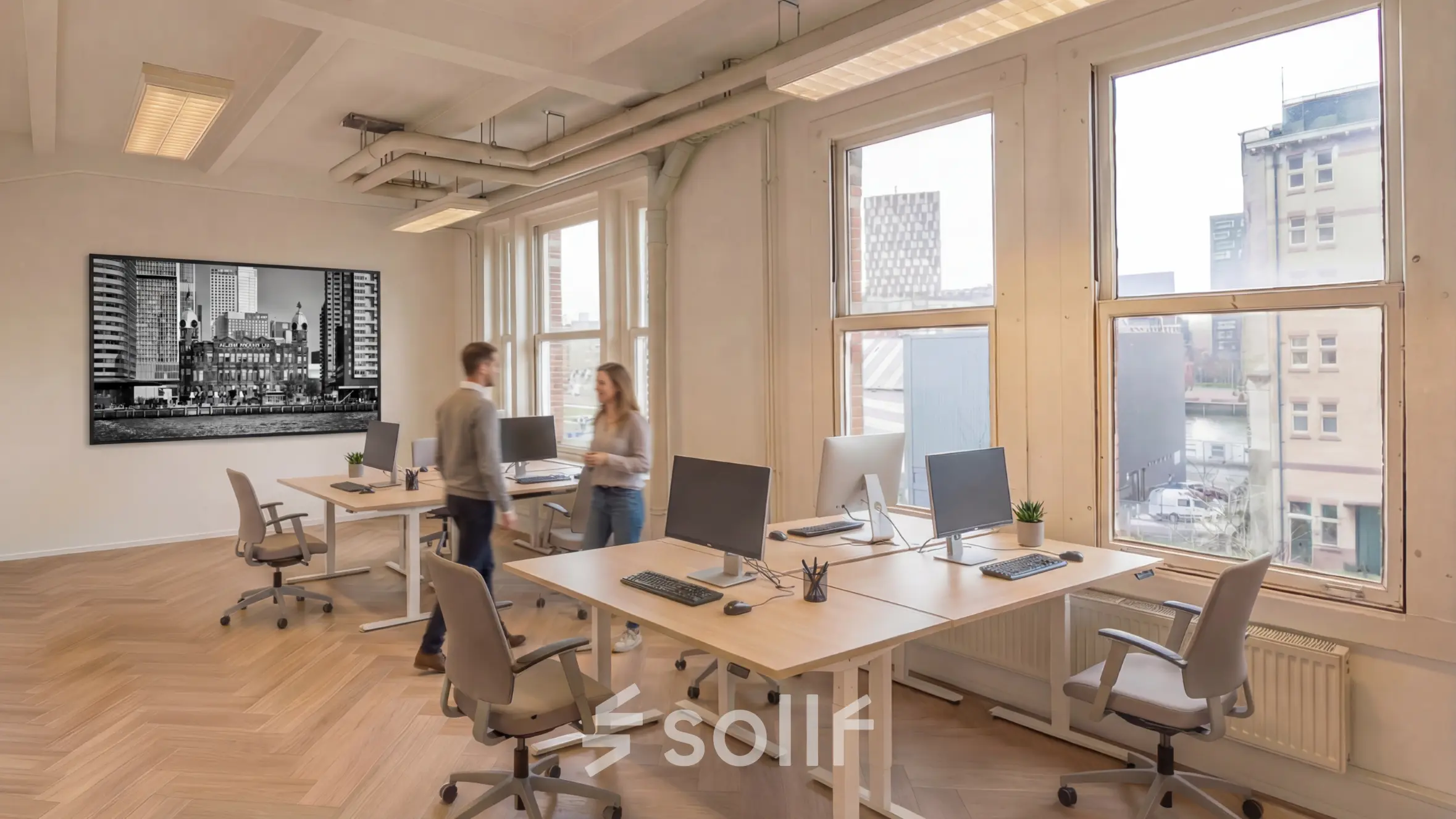 Lichte en ruime kantoor met houten vloer, grote ramen en bureaus met computers waar twee personen met elkaar werken of overleggen.