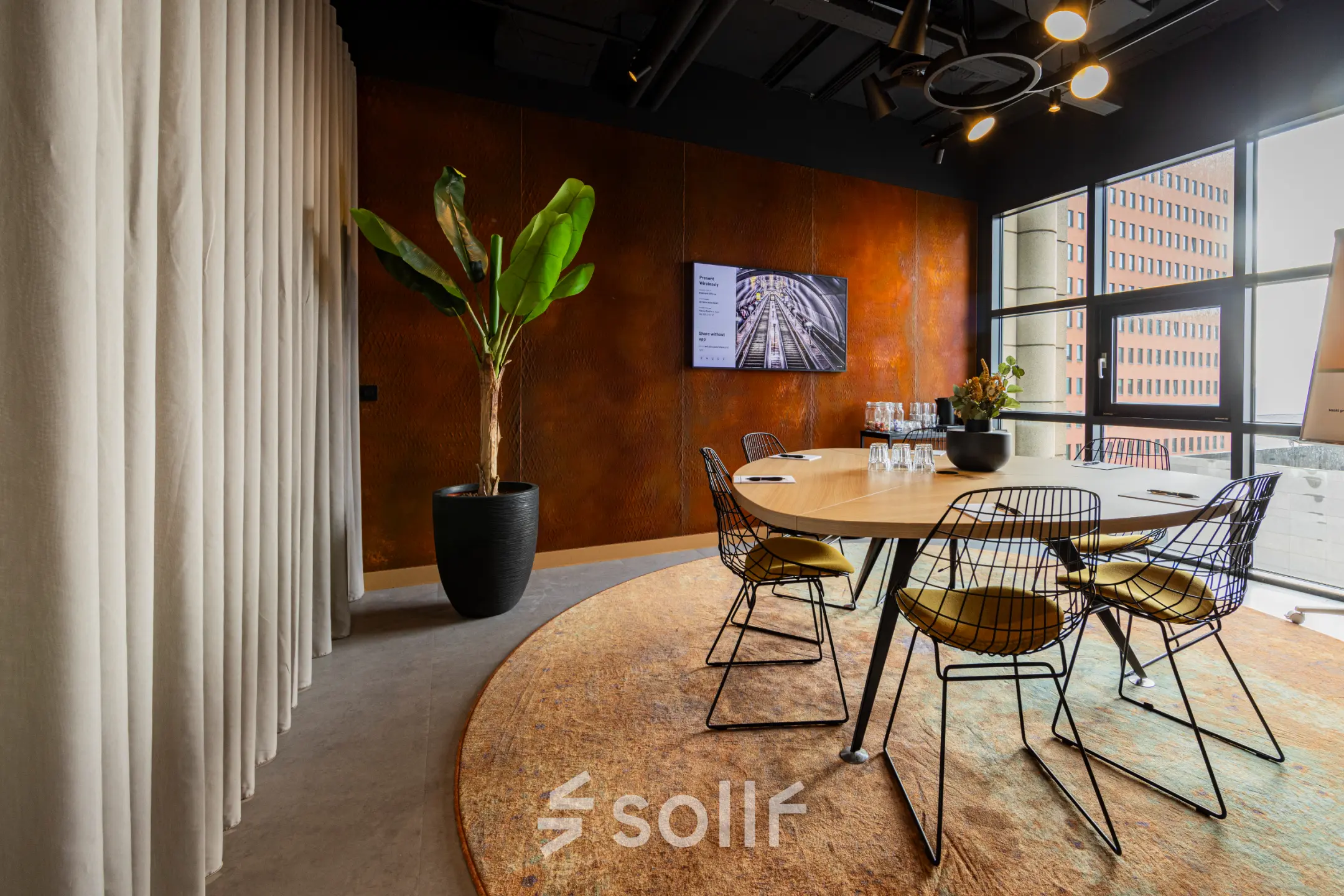 Moderne vergaderruimte met grote ramen, ronde tafel en een grote plant, gelegen op Wilhelminaplein 1-40, Rotterdam Kop van Zuid. Ideaal voor kantoorruimte huren en zakelijke bijeenkomsten.