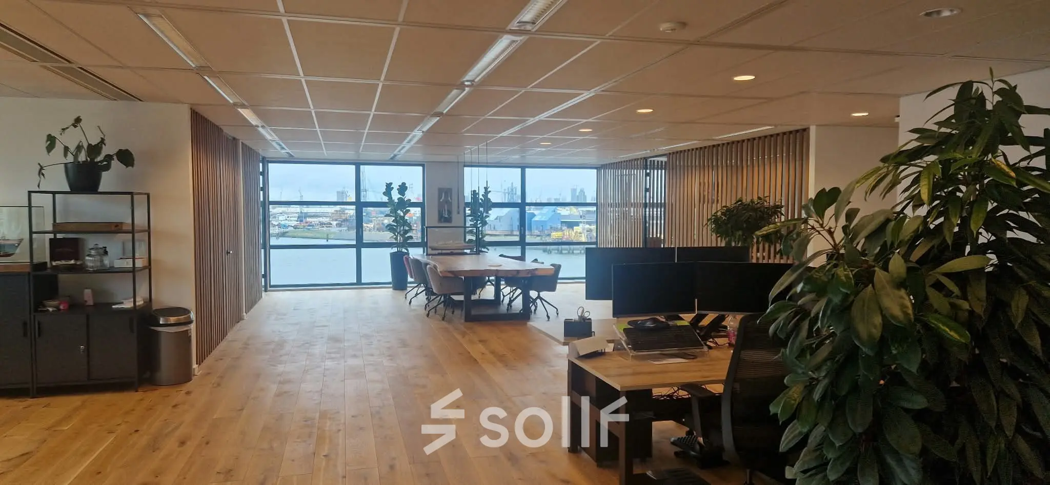 Ruime kantoorruimte met houten vloer en uitzicht op de haven. De ruimte heeft werkplekken, planten en biedt diverse zitmogelijkheden. Ideaal voor bedrijven die kantoorruimte willen huren in Rotterdam Waalhaven.