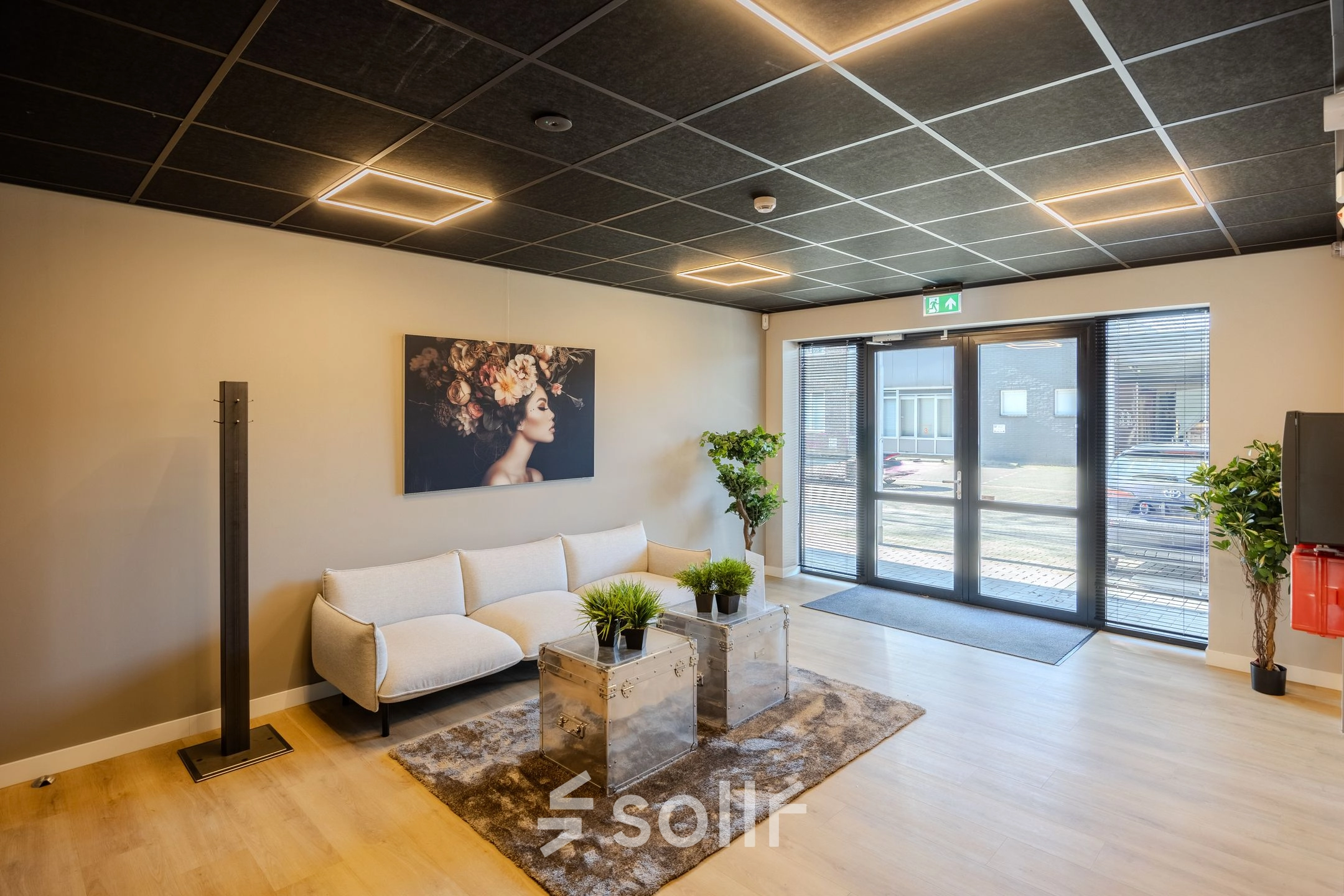 Moderne ontvangstruimte in een kantoorpand met een stijlvolle witte bank, glazen salontafel en kunst aan de muur. Gezellige verlichting en groene planten verfraaien de ruimte. Ideaal voor kantoorruimte huren.