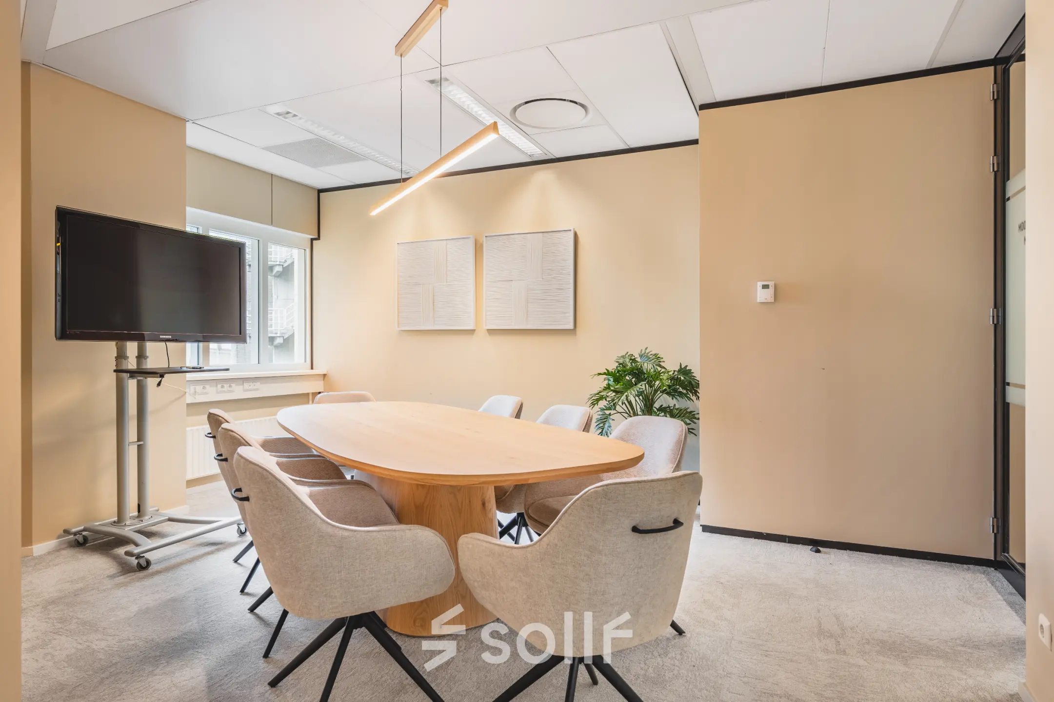 Vergaderruimte in kantoorpand te Utrecht Centraal Station met ovale houten tafel, acht stoelen, en een groot scherm. Neutrale kleuren en natuurlijke verlichting geven een zakelijke sfeer.