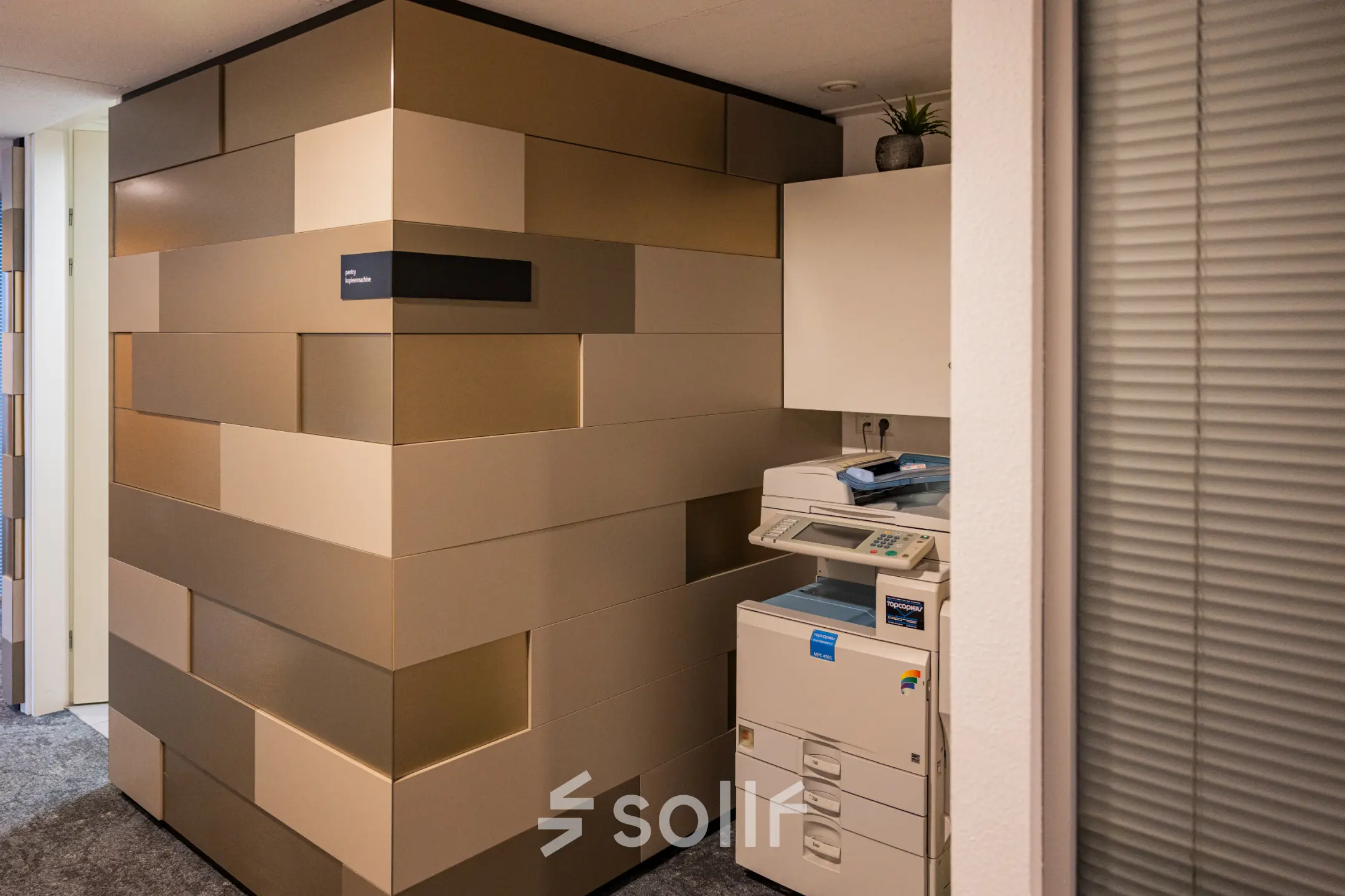 Binnen een kantoorpand op Middenburcht 136, Utrecht Leidsche Rijn staat een kopieerapparaat naast een modern ontworpen muur. Ideale plek voor bedrijven om hun dagelijkse taken efficiënt uit te voeren.