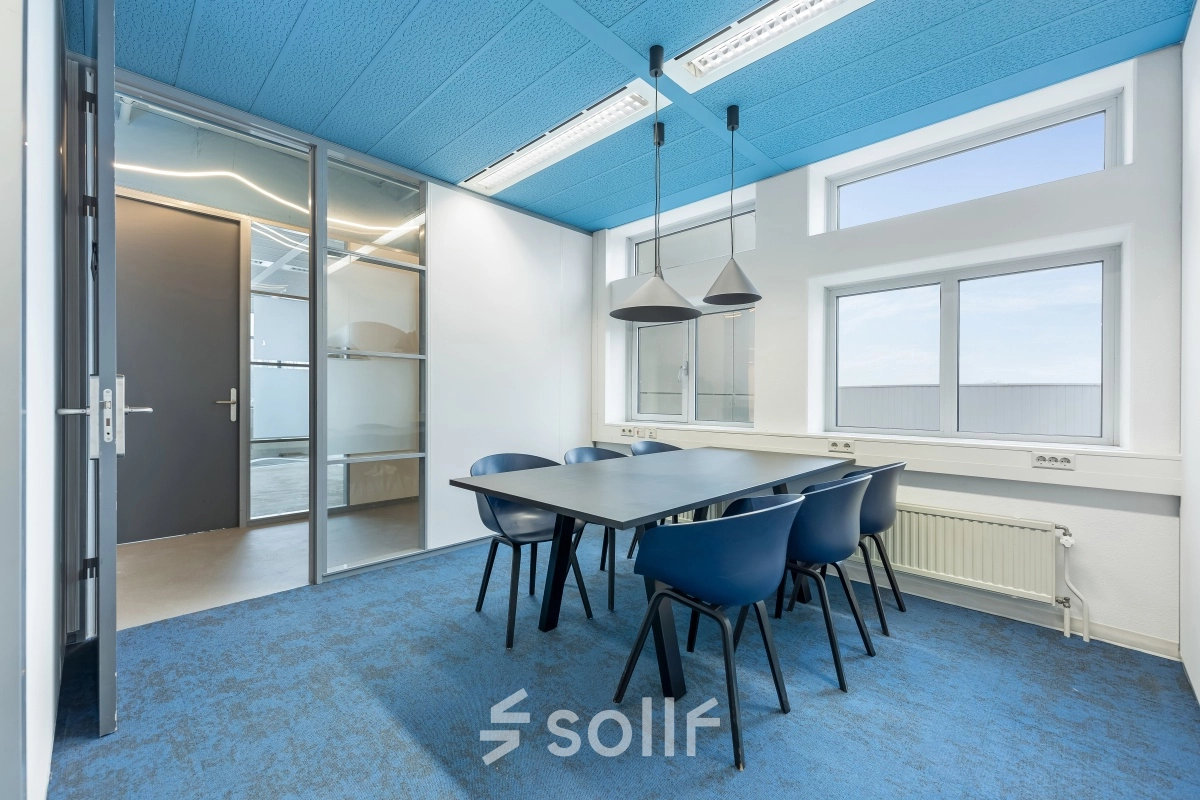 Moderne, goed verlichte kantoorruimte met blauwe accenten, voorzien van een tafel en zes stoelen, ideaal voor vergaderingen of als werkruimte. Grote ramen bieden veel natuurlijk lichtinzichten. Geschikt voor kantoorpand huren.