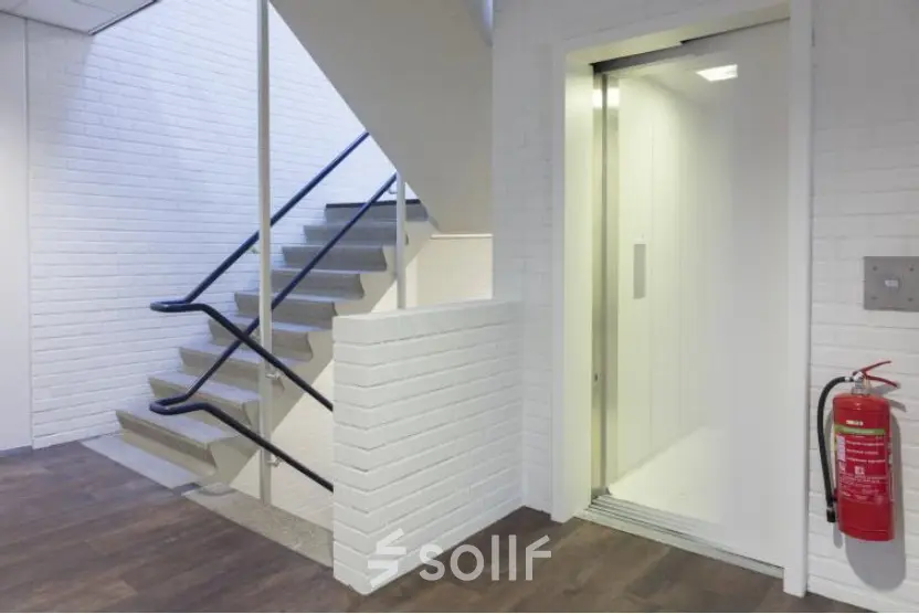Ingang van een kantoorpand op Pompmolenlaan 9, Woerden, met een open lift en trap. Ideaal voor wie kantoorruimte wil huren. Heldere verlichting en praktische indeling creëren een professionele uitstraling.