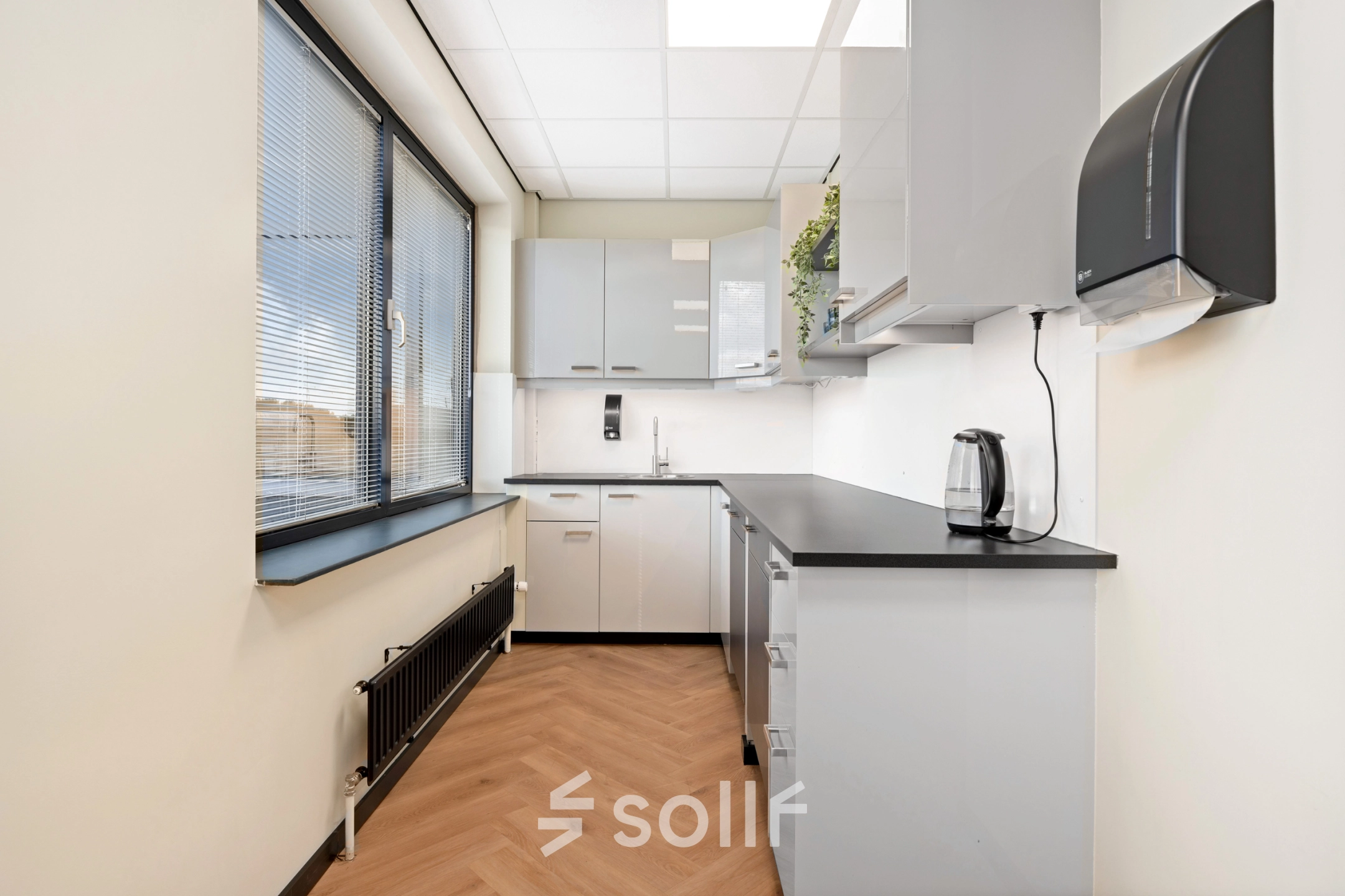 Moderne pantry in kantoorpand op Stationsplein 4, Zwijndrecht, voorzien van strakke keukenkasten, een waterkoker en een groot raam. Perfecte locatie om kantoorruimte te huren. Functioneel en uitnodigend ontwerp.