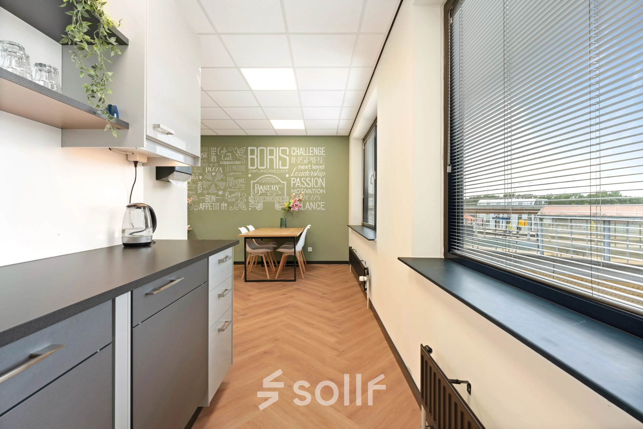 Kantoorruimte huren op Stationsplein 4, Zwijndrecht. Moderne pantry met lichtinval via grote ramen, uitgerust met keukenkastjes, beklede vloer en stoelen. Ideaal voor zakelijke pauzes en informele vergaderingen.