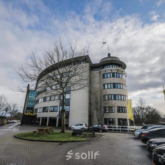 Het ronde kantoorpand op Hanzelaan 351-361 in Zwolle met parkeerplaatsen en een blauwe lucht op de achtergrond.