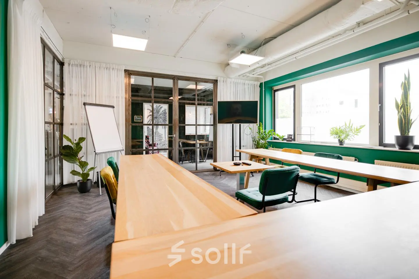 Kantoorruimte huren op Hanzeplein 11-27 in Zwolle met houten tafels, een flip-over en een groot scherm. Groen-witte tinten met veel lichtinval, ideaal voor vergaderingen. Planten voegen een frisse sfeer toe.