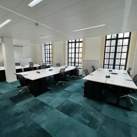 Ruime kantoorruimte te huur op Rue des Colonies 56, Brussel Europese Wijk, met grote ramen en blauwe vloertapijten. Verschillende bureaus en stoelen klaar voor gebruik in een lichte omgeving.
