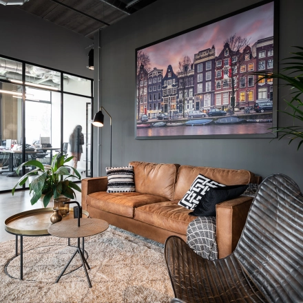 Kantoor te huur in Leuven met moderne, gemeubileerde zithoek, inclusief een bruine leren bank, bijzettafels en groene planten. Op de achtergrond zijn mensen aan het werk in een glazen ruimte.