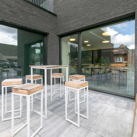 Stijlvolle buitenruimte met moderne hoge tafels en krukken, ideaal voor informeel overleg. Grote ramen bieden uitzicht op kantoorruimte te huur bij Olenseweg 375 in Westerlo.