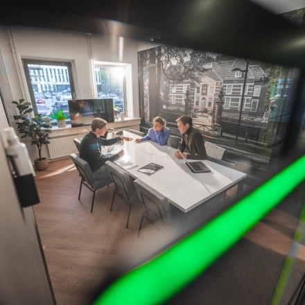 Modern office space featuring a large table with three people engaged in a business meeting, equipped with tablets and documents. Bright, professional setting ideal for office space rental.