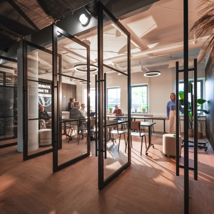 Modern office space rental at Egbert Gorterstraat 2-14, Almelo, featuring glass-walled work areas with people engaged in business discussions under stylish lighting.
