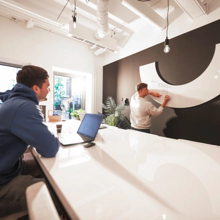 Modern office space rental in Almelo with a sleek interior, featuring a collaborative work environment. Two professionals engage in brainstorming at a high gloss table with a stylish black and white backdrop.
