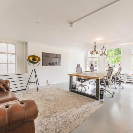 Bright office space for rent at Herengracht 221, Amsterdam Canal Belt, with modern furnishings, including a large wooden table and chairs, facing large windows.