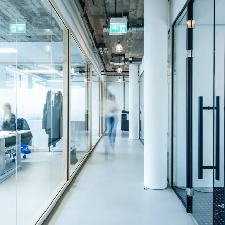 Modern office space rental at Singel 542, Amsterdam Canal Belt. Glass partitions and visible ceiling add a contemporary touch. Individuals are engaged in business activities.