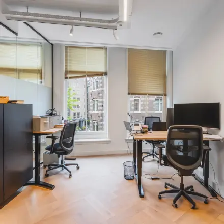 Bright office space with wooden floors, desks, chairs, and a large window at Hekelveld 8-10, Amsterdam Center. Ideal for office space rental.