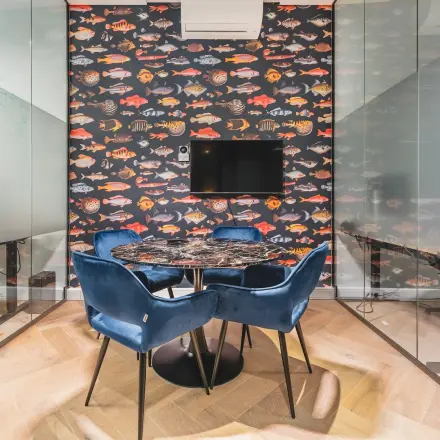 Bright workspace in Amsterdam Center with a stylish fish-patterned wall, glass partitions, and a modern round table surrounded by blue chairs for rent.