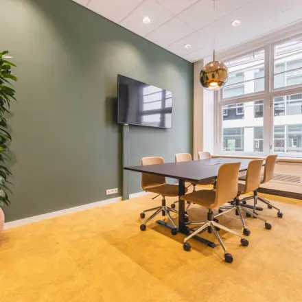 Bright office space for rent at Herengracht 471, Amsterdam Center, featuring a meeting table, chairs, wall-mounted TV, and large window with natural light.