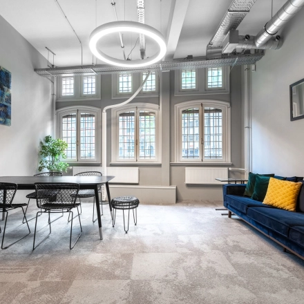 Bright office space for rent at Nieuwezijds Voorburgwal 104-108 in Amsterdam Center, featuring a modern design with a table, chairs, and a sofa under large, arched windows.
