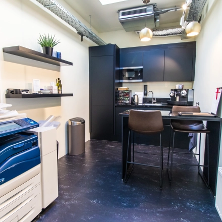 Compact, furnished kitchenette in an Amsterdam office, featuring a high table with chairs, appliances, and a printer. Ideal for small office space rental.