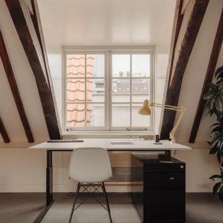 Cozy office space for rent at Prins Hendrikkade 14, Amsterdam Center, featuring a single desk with a lamp and drawers under rustic wooden beams.